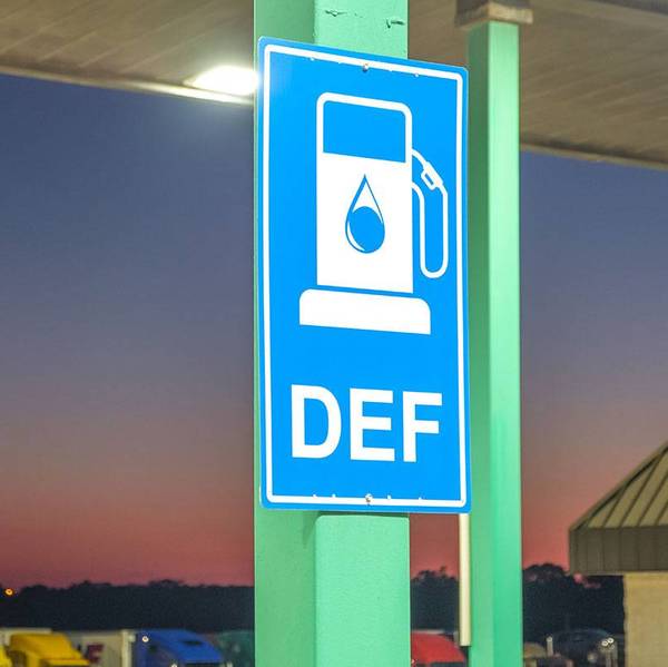 A blue DEF sign hanging on a green post at a fuel station.