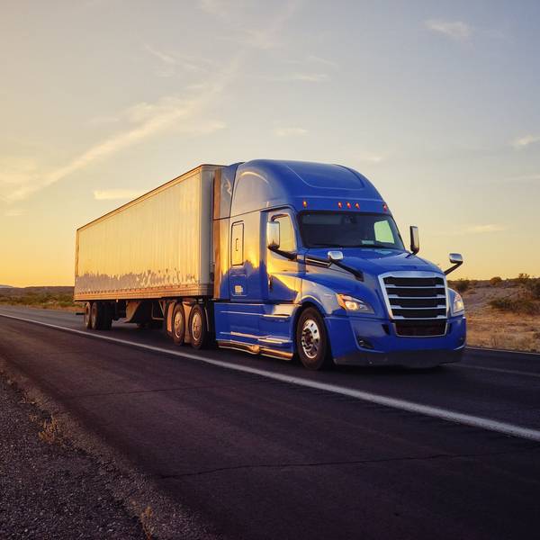 A blue semi-truck drives down a road at sunset.