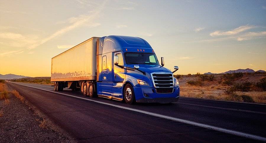 A blue semi-truck drives down a road at sunset.