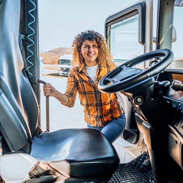 A female truck driver climbs into the cab of a semi truck while holding onto hand rails.