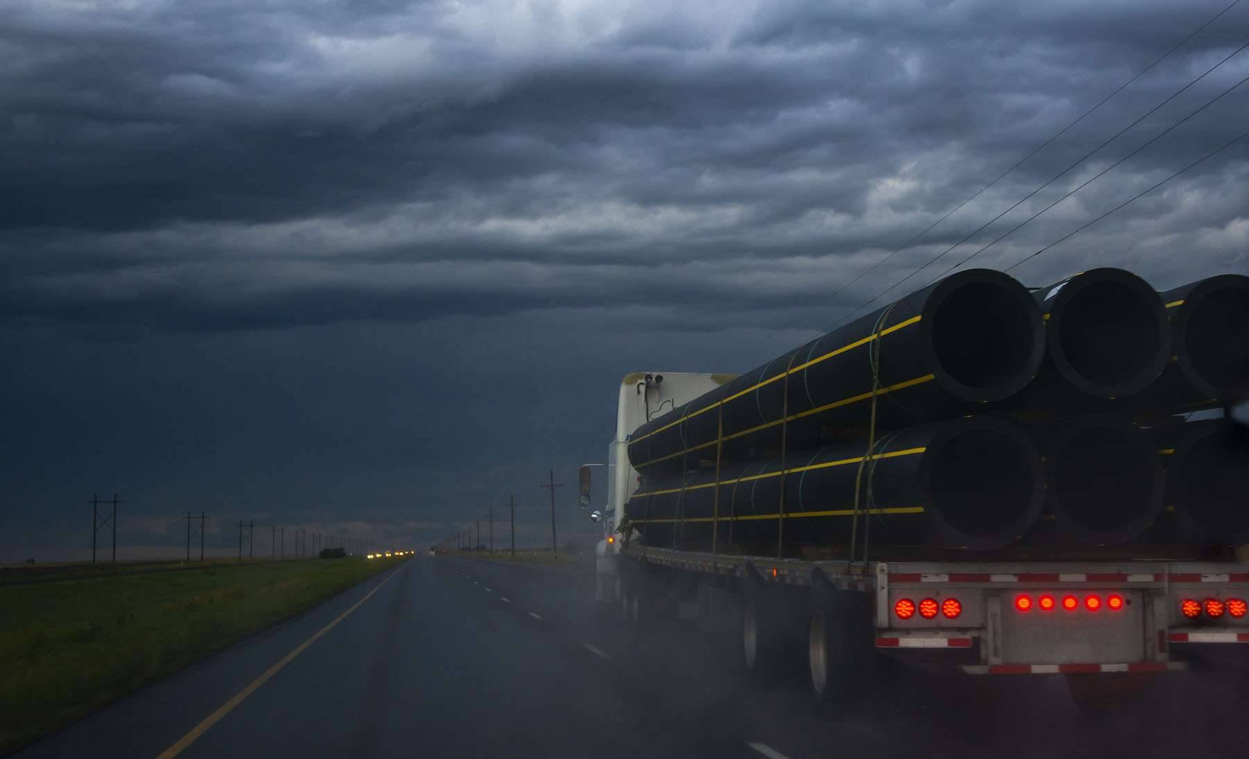 A flatbed semi-truck carrying pipes drives under a stormy sky.