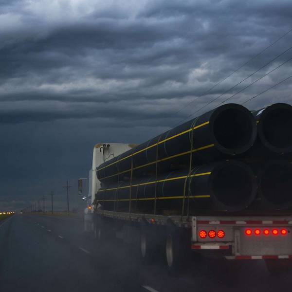 A flatbed semi-truck carrying pipes drives under a stormy sky.