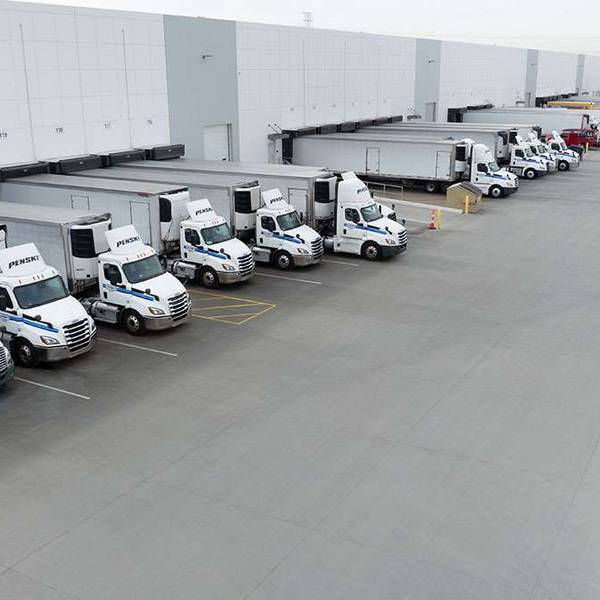 A fleet of white Penske semi-trucks are parked in a line backed up to a big warehouse loading dock.