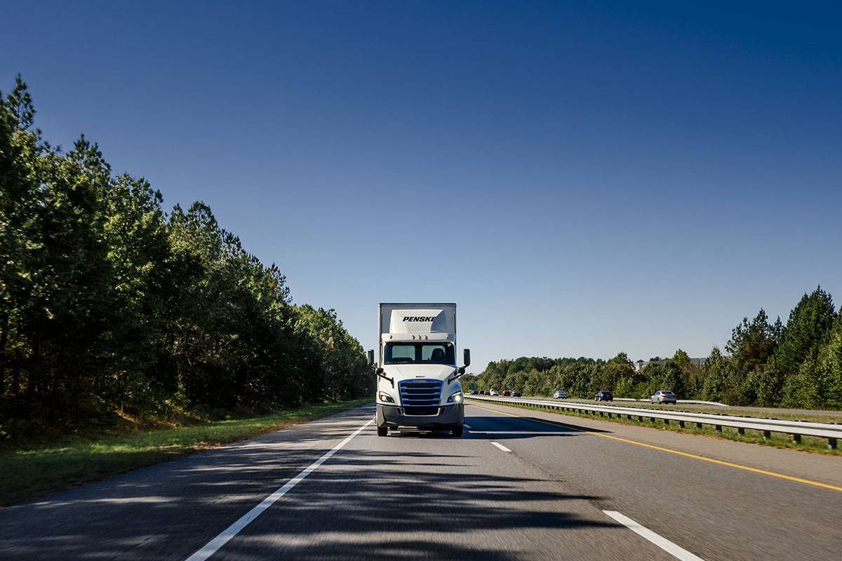 A front shot of a white Penske semi-truck driving towards the camera under a blue sky.