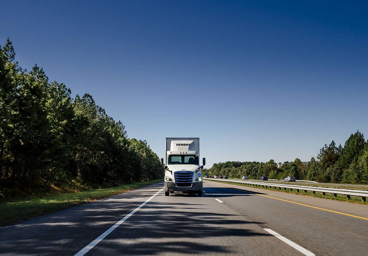 A front shot of a white Penske semi-truck driving towards the camera under a blue sky.