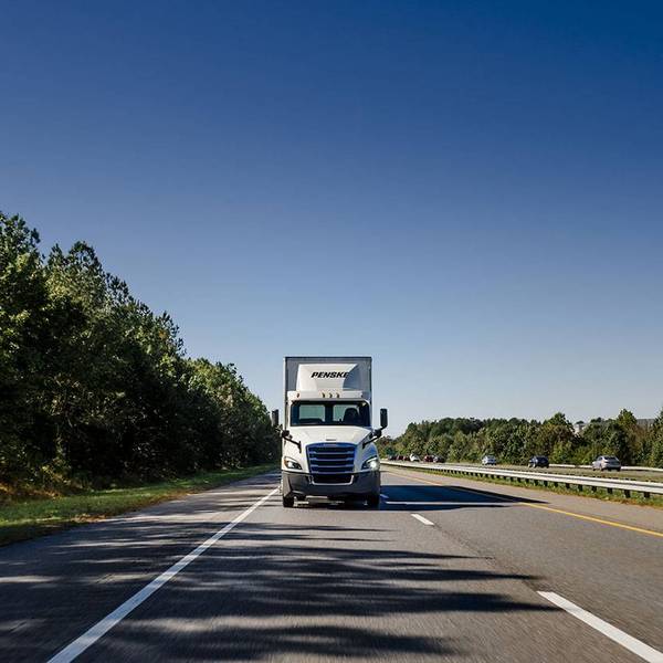 A front shot of a white Penske semi-truck driving towards the camera under a blue sky.