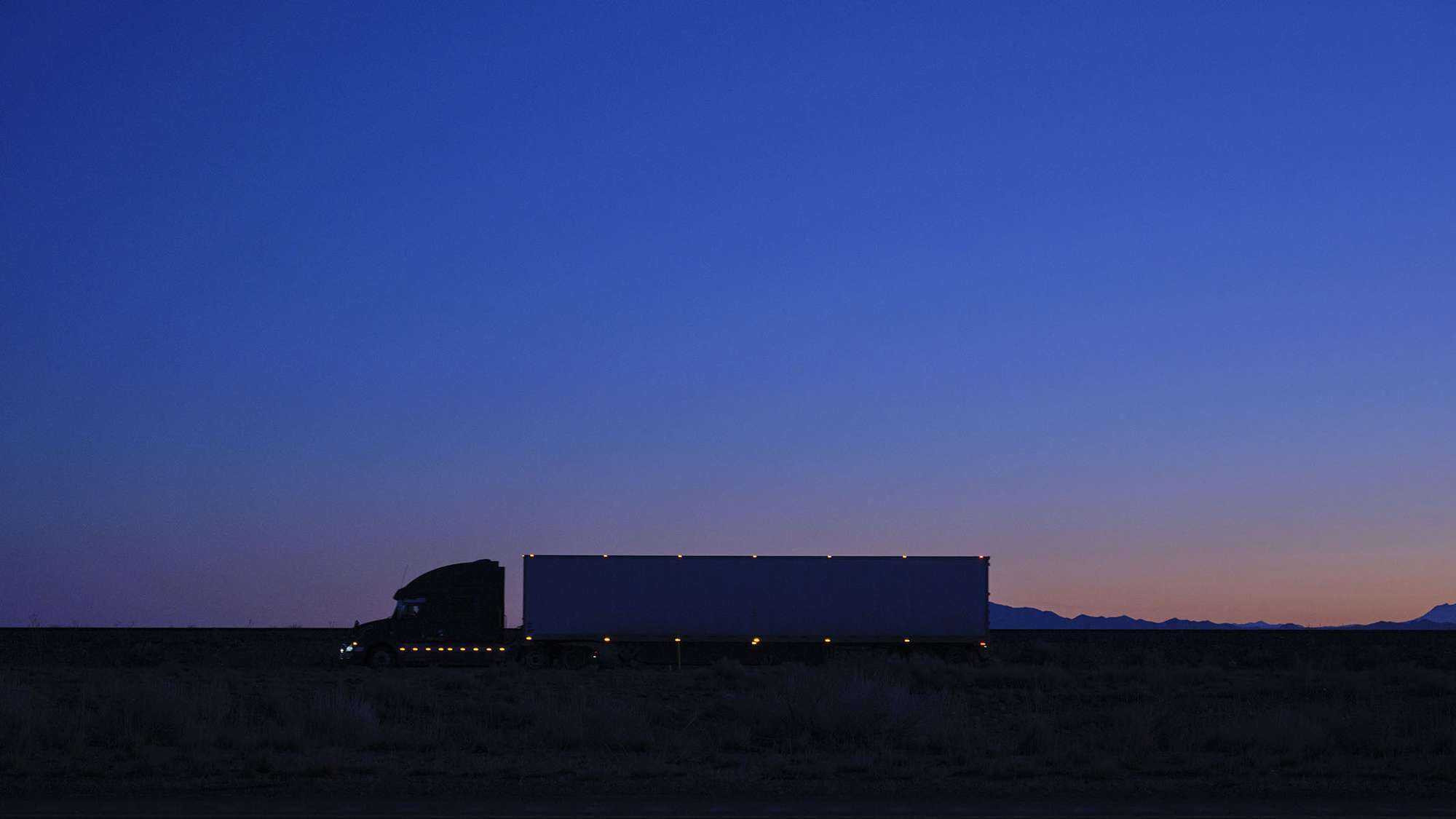 A full shot of a semi-truck with lights driving at dusk.
