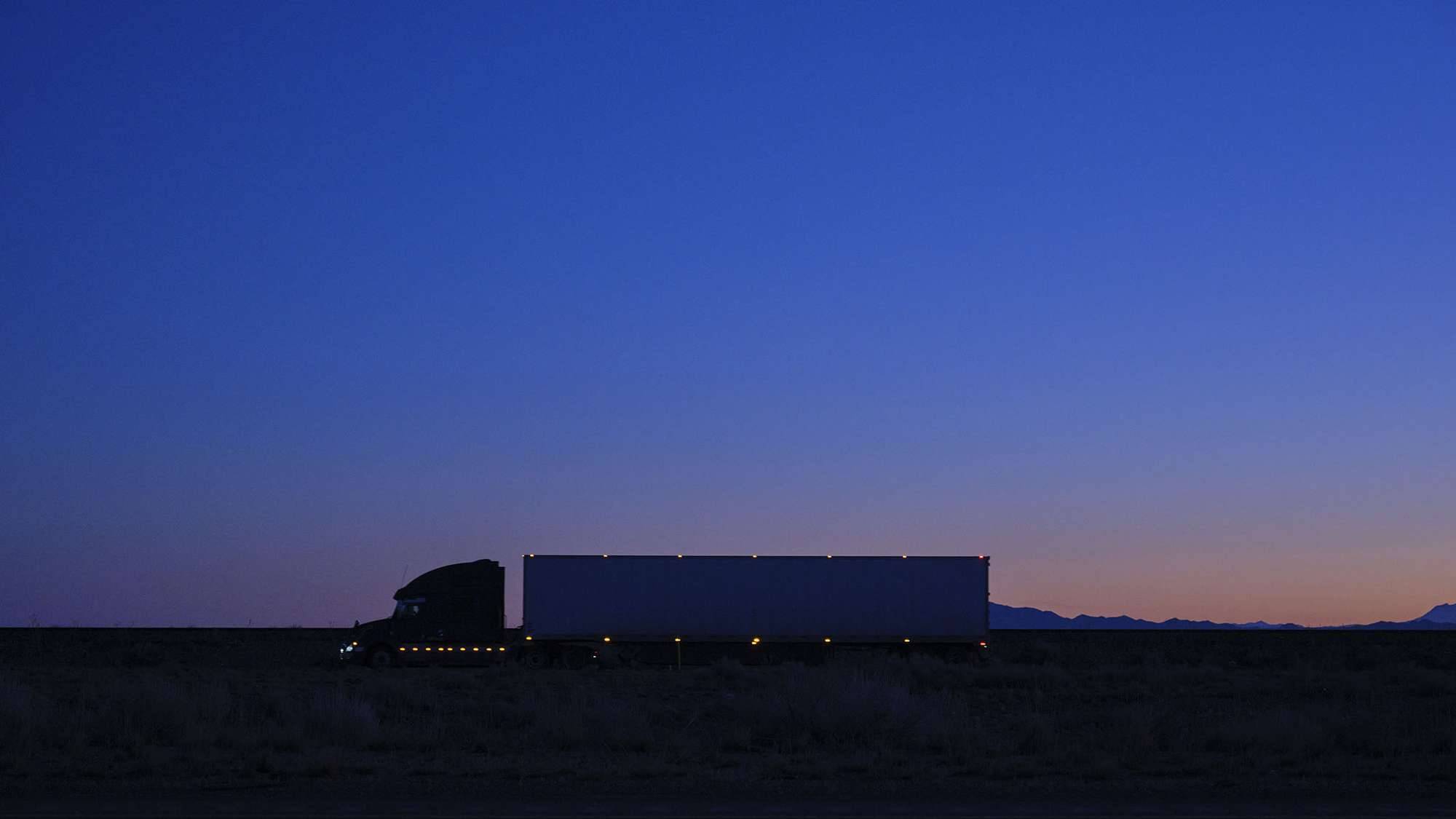 A full shot of a semi-truck with lights driving at dusk.