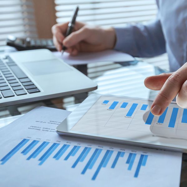 A hand points to a blue and white bar chart while the other hand holds a pen and takes notes by a laptop keyboard.