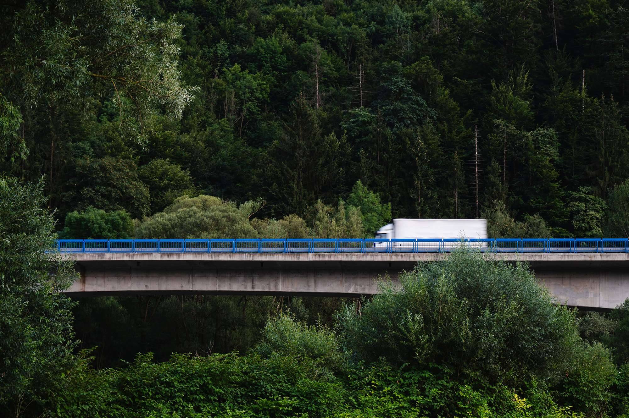 A large white box truck drives over a bridge through a lush green forest.