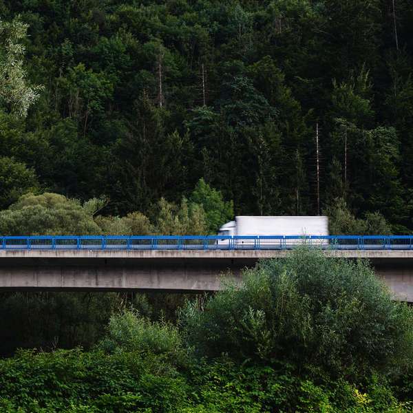 A large white box truck drives over a bridge through a lush green forest.