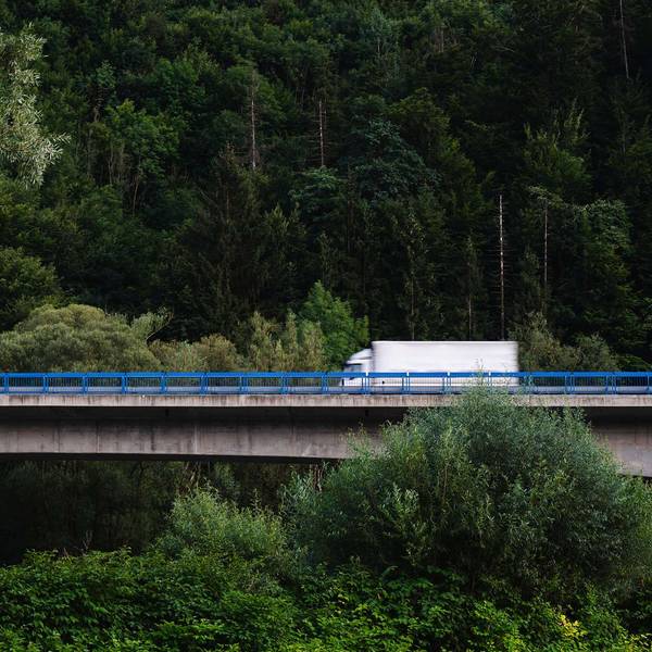 A large white box truck drives over a bridge through a lush green forest.