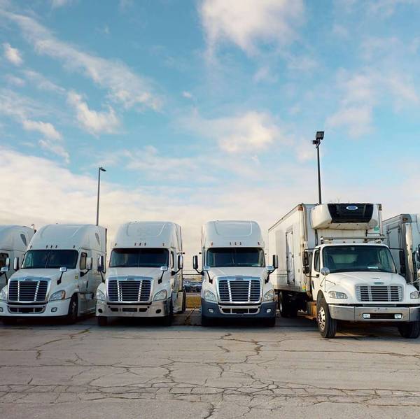 A line of white semi-trucks and box trucks parked in a lot against a blue cloudy sky.