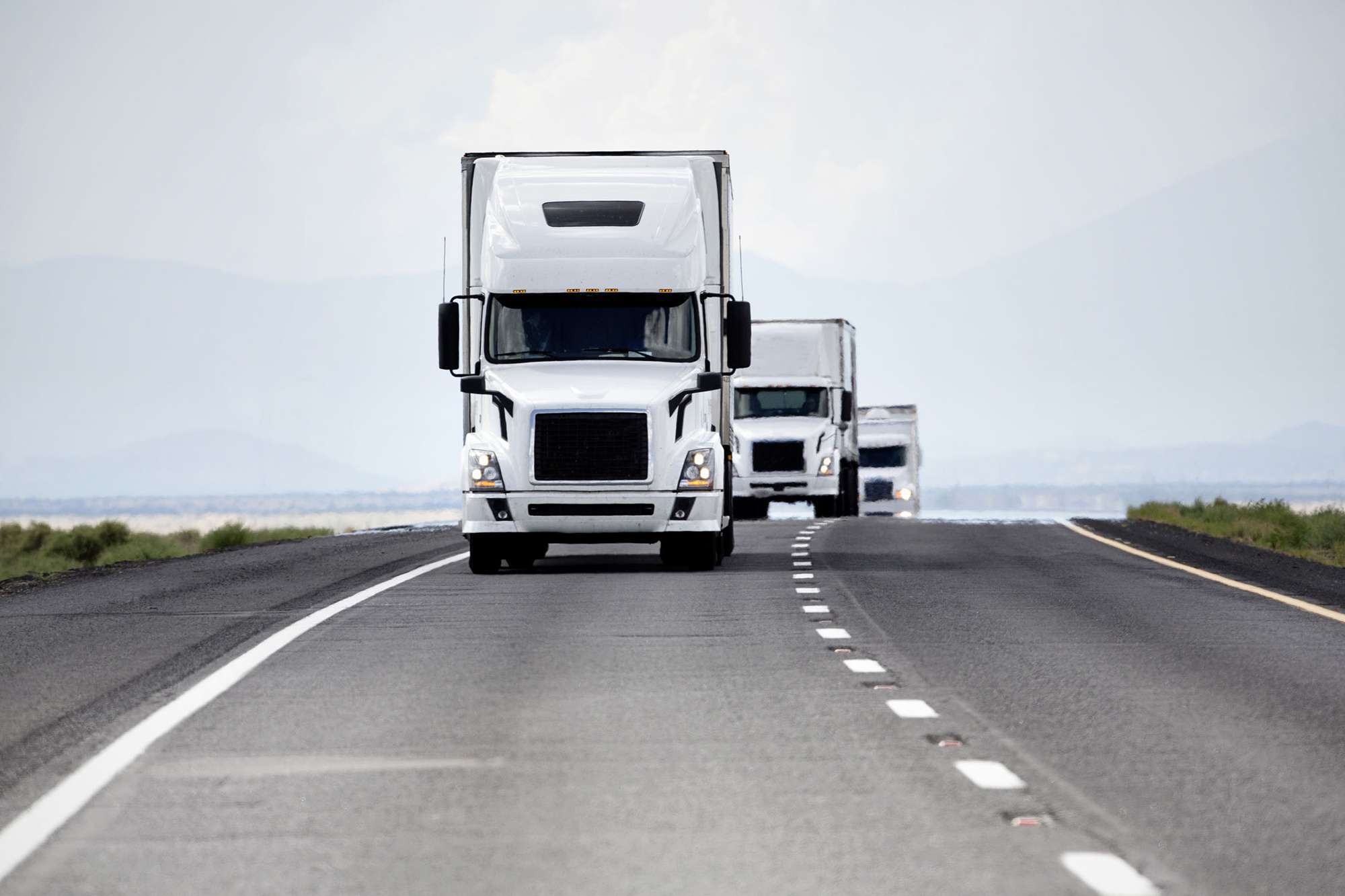 A line of white semi-trucks drive up the road.