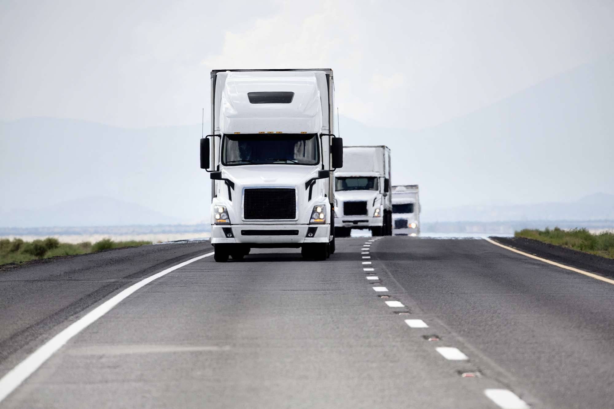 A line of white semi-trucks drive up the road.