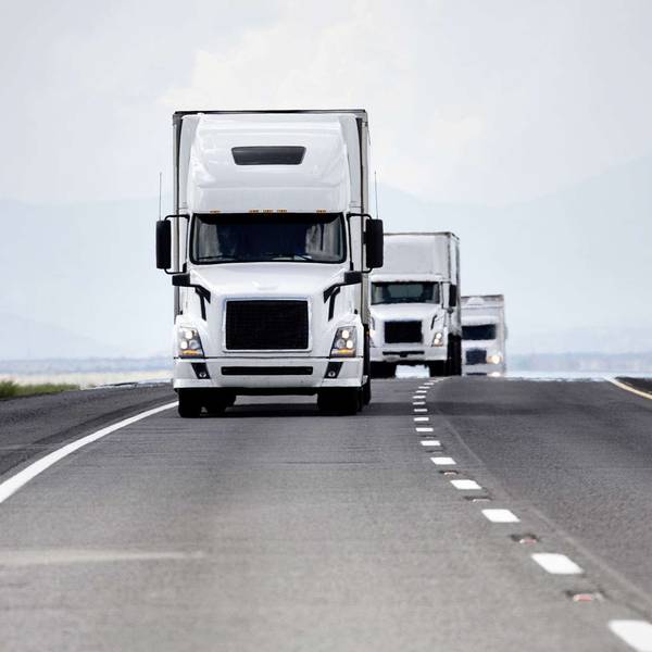A line of white semi-trucks drive up the road.