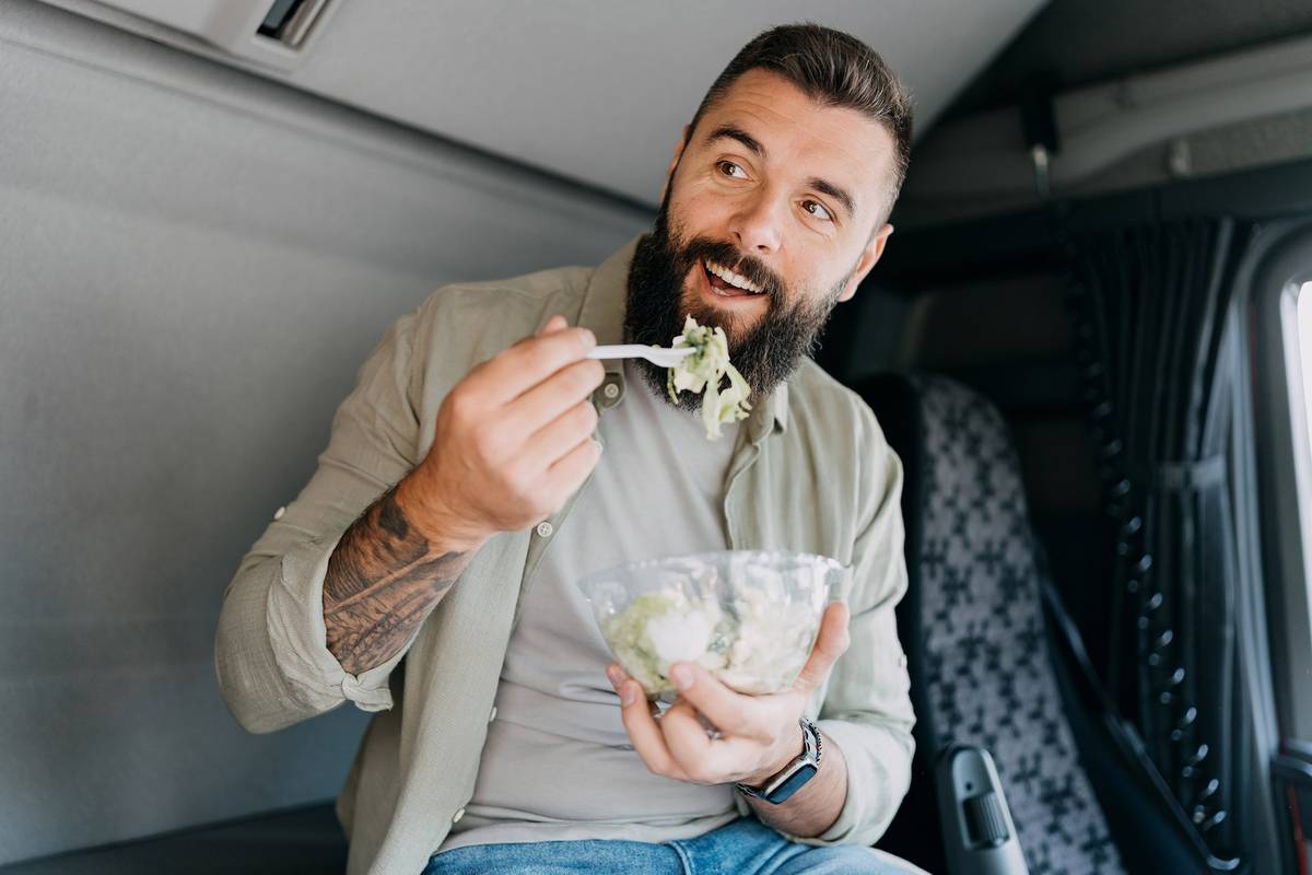 A man eats a salad while sitting in the cab of a semi-truck.
