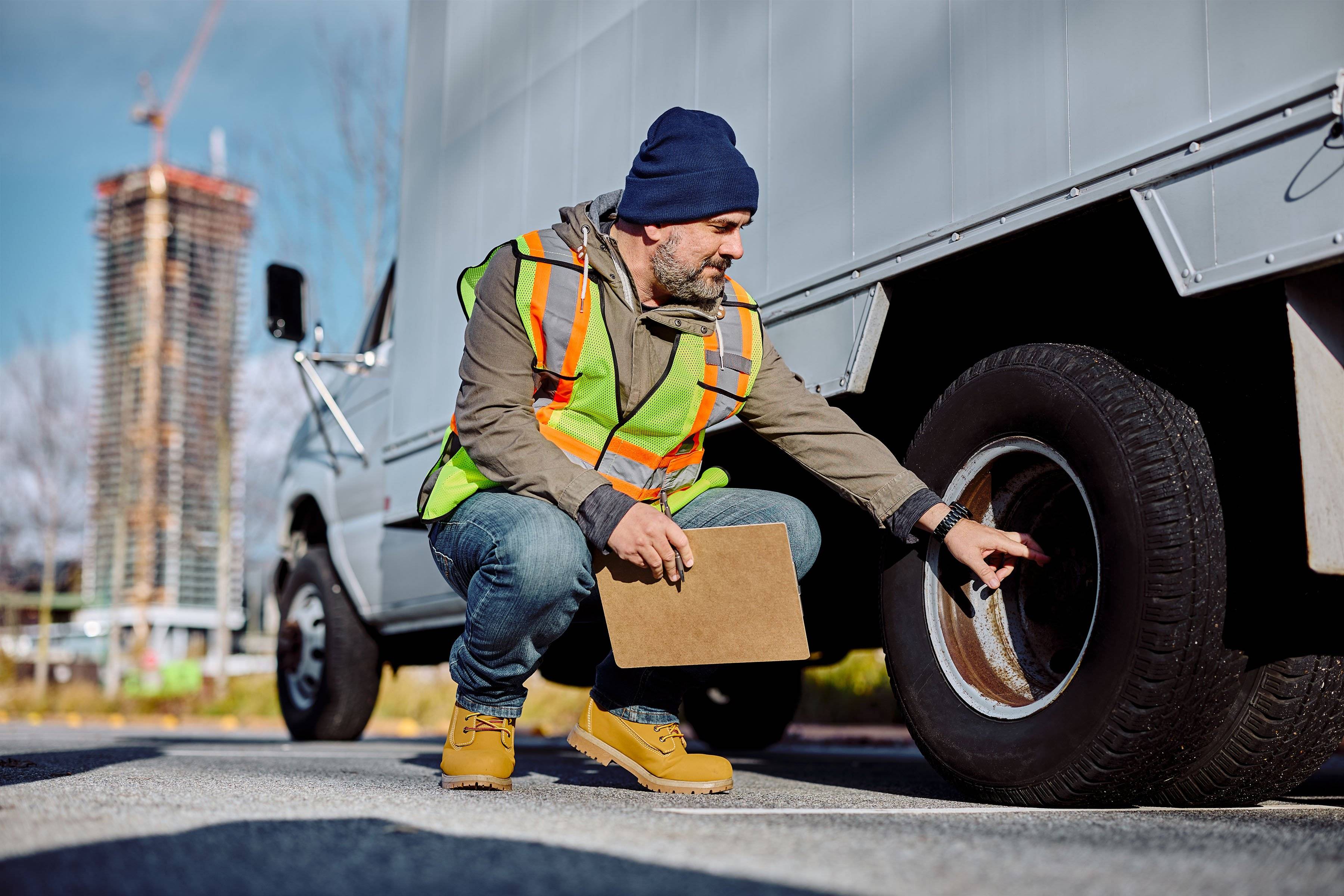 A man holding a clipboard squats down to check a box truck tire.