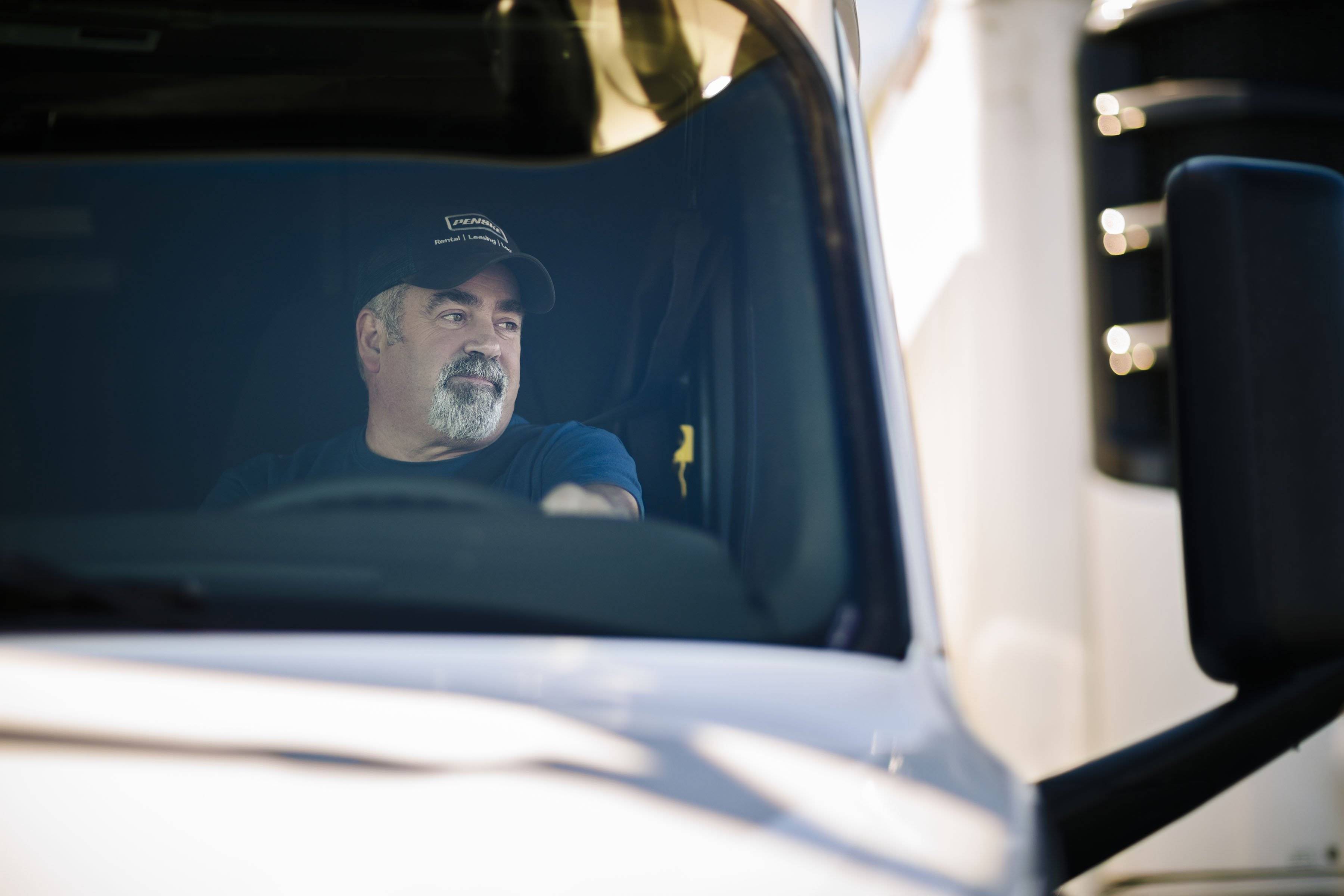 A man in a blue Penske hat sitting behind the wheel of a white truck.