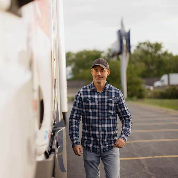 A man in a blue plaid flannel shirt walks in a parking lot next to a white semi-truck.