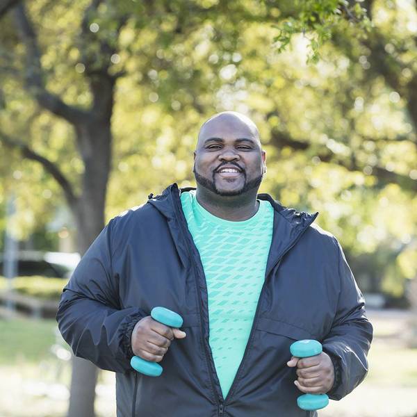 A man in a t-shirt and light jacket walks with blue handweights in his hands.