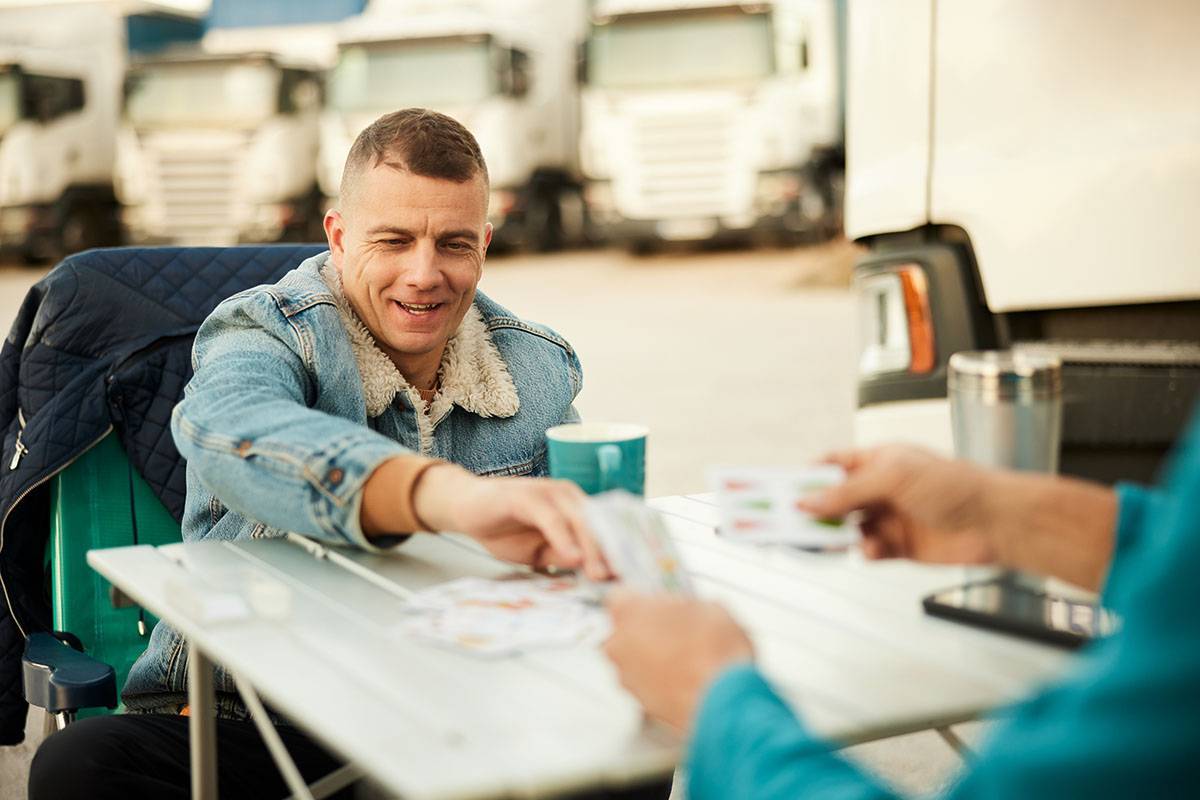 A man sits in a semi-truck parking lot playing cards with another person.
