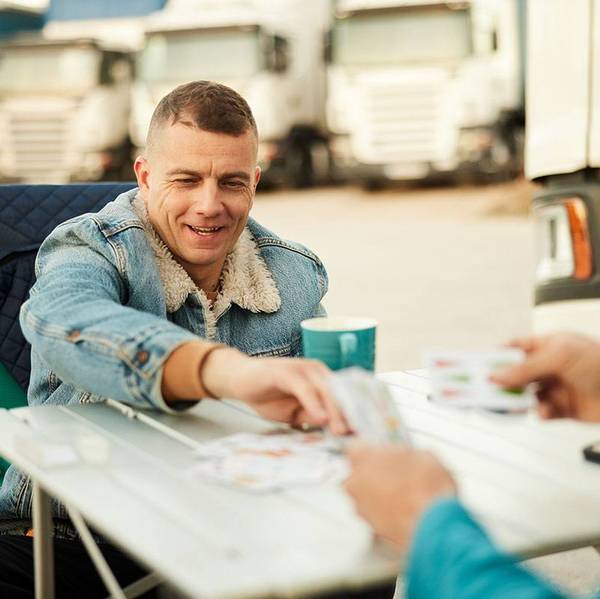 A man sits in a semi-truck parking lot playing cards with another person.