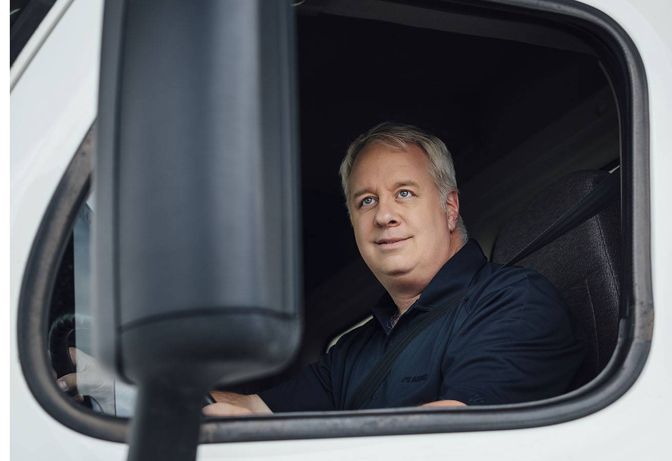 A man sits in the driver seat of a white truck looking out the window.