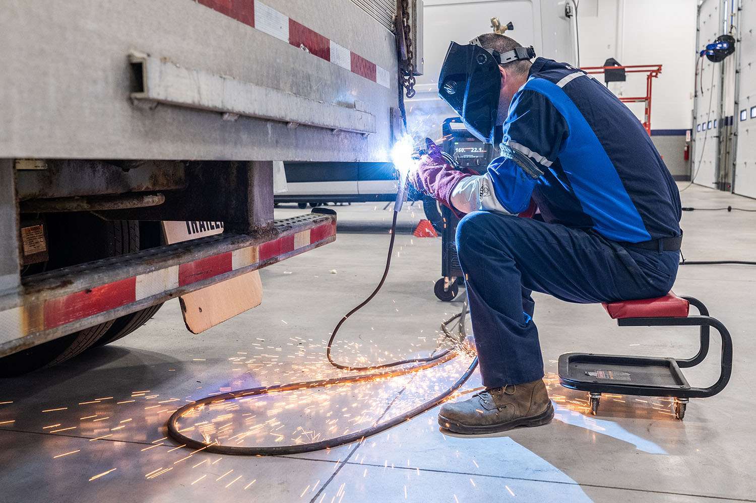 A man sits on a stool and sauders something on the back end of a semi-truck trailer.