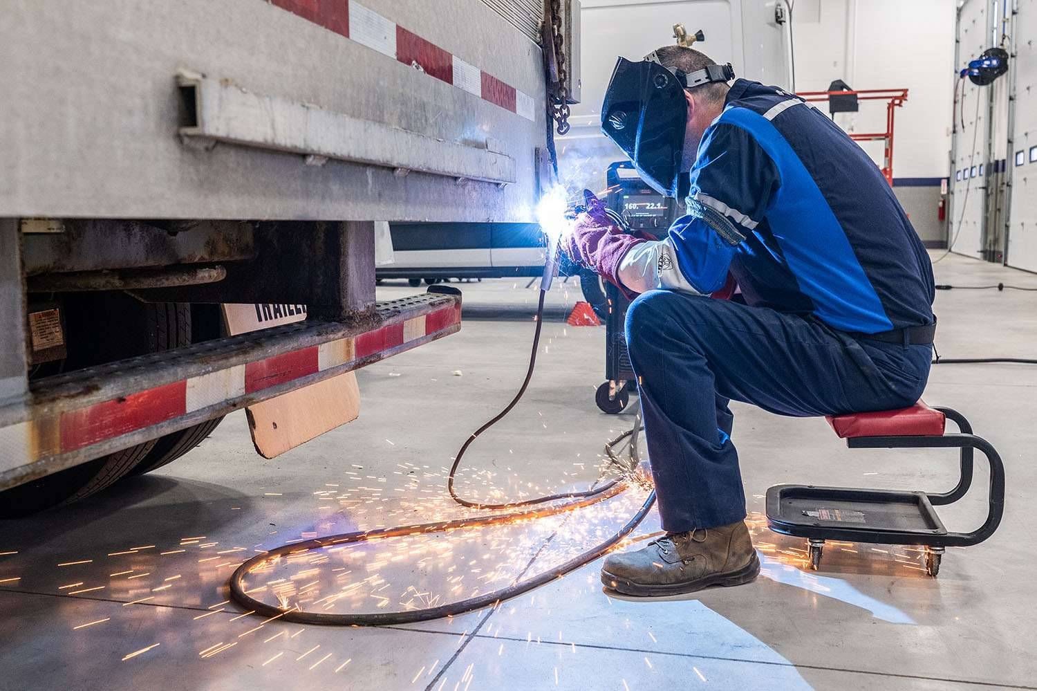 A man sits on a stool and sauders something on the back end of a semi-truck trailer.