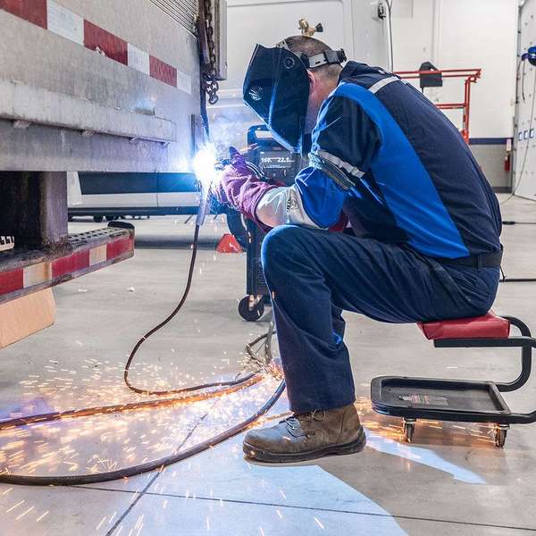 A man sits on a stool and sauders something on the back end of a semi-truck trailer.