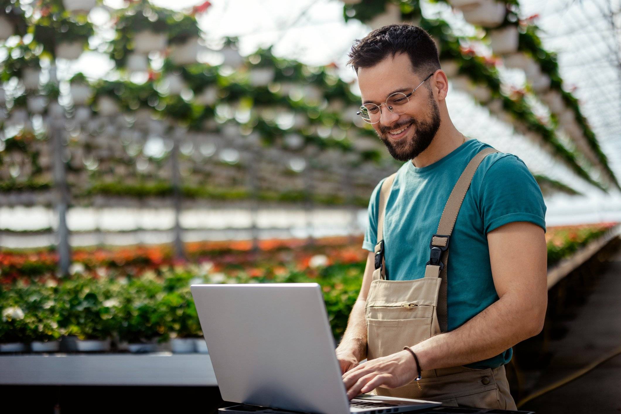 A man stands in a greenhouse working on his laptop.