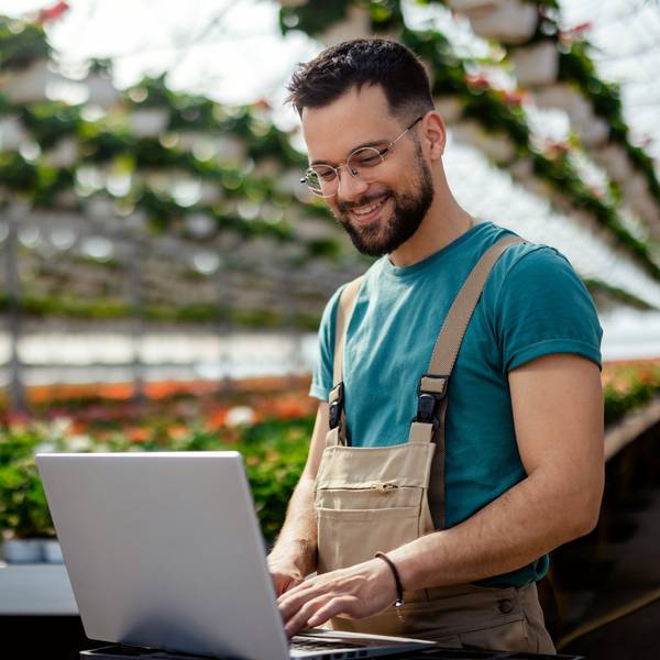 A man stands in a greenhouse working on his laptop.