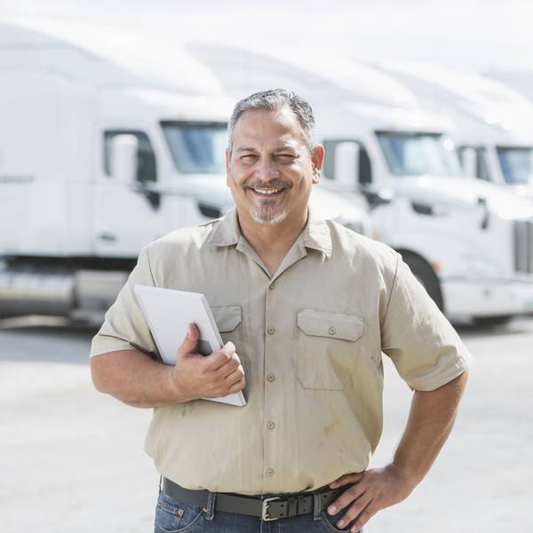 A man with a grey beard stands with his hand on his hip in front of a fleet of white semi-trucks.
