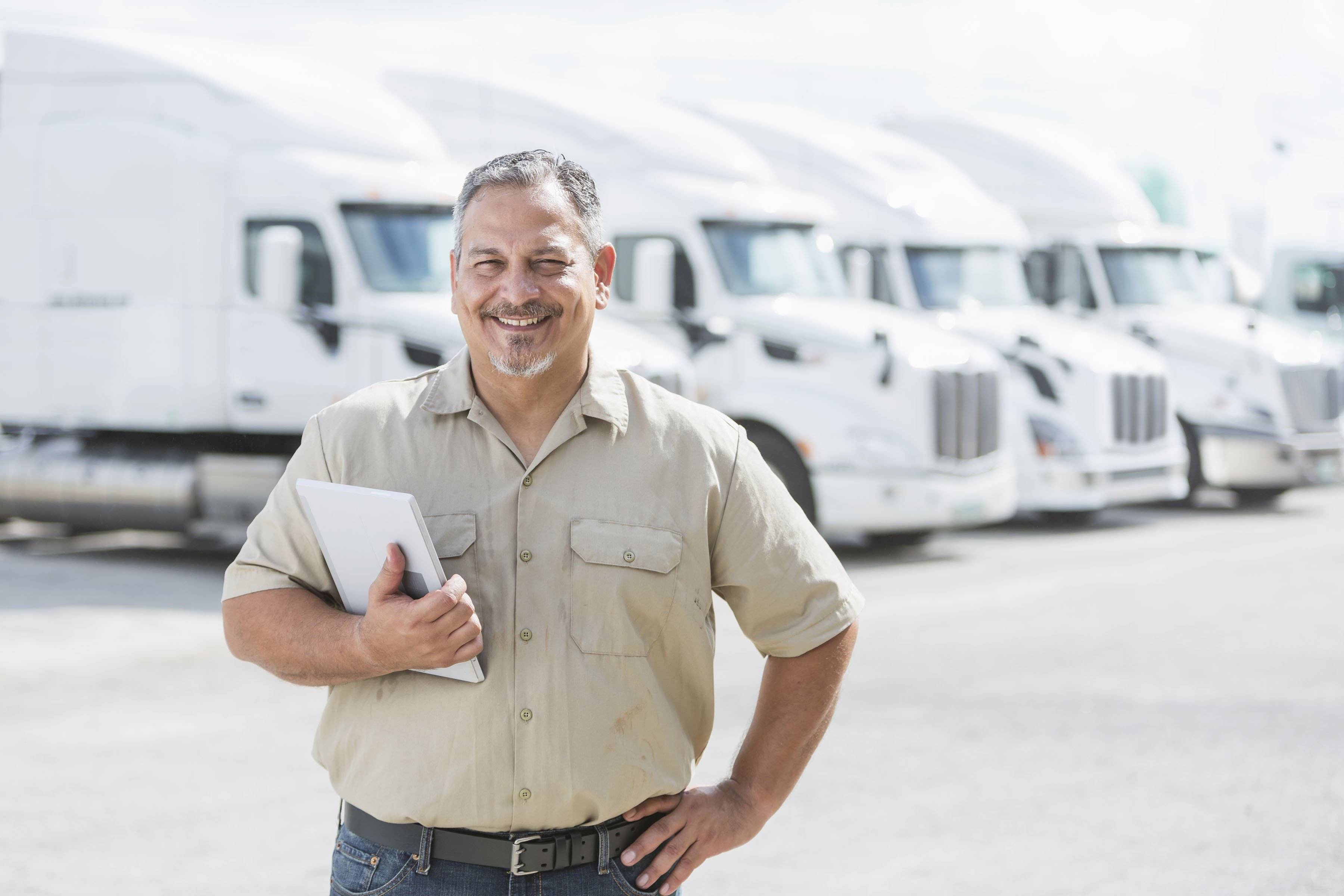 A man with a grey beard stands with his hand on his hip in front of a fleet of white semi-trucks.