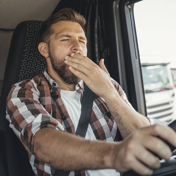 A man with a plaid shirt sits in a truck cab and covers his mouth with his hand while he yawns.
