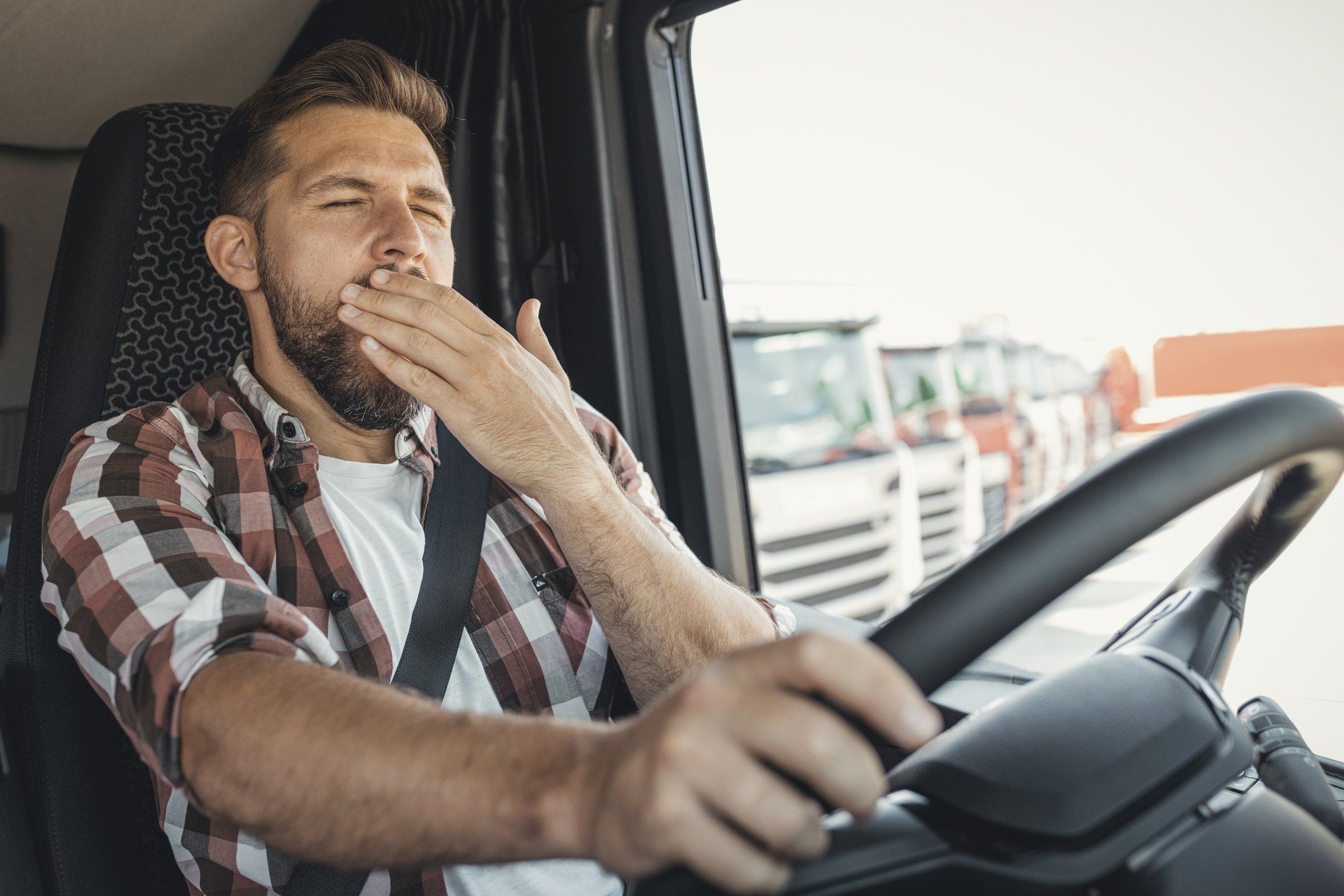 A man with a plaid shirt sits in a truck cab and covers his mouth with his hand while he yawns.