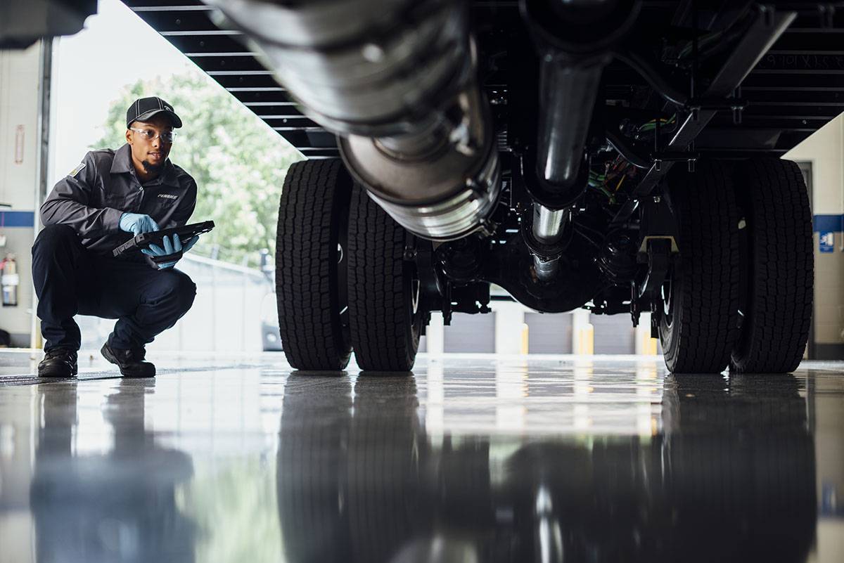 A Penske technician holding a tablet kneels down to look under a semi-truck.