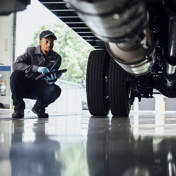 A Penske technician holding a tablet kneels down to look under a semi-truck.
