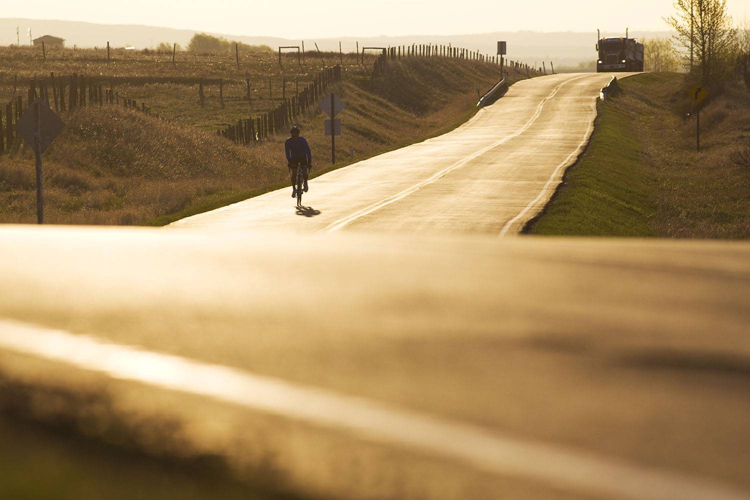 A person rides a bike down a road at sunset with a semi-truck behind.