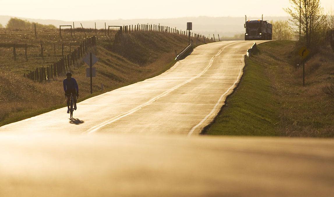 A person rides a bike down a road at sunset with a semi-truck behind.