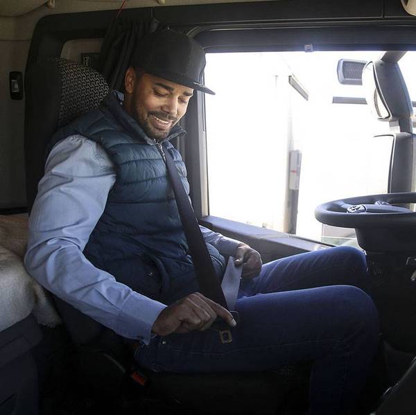 A truck driver in a blue vest and hat buckles his seat belt while sitting in the cab of a semi-truck.