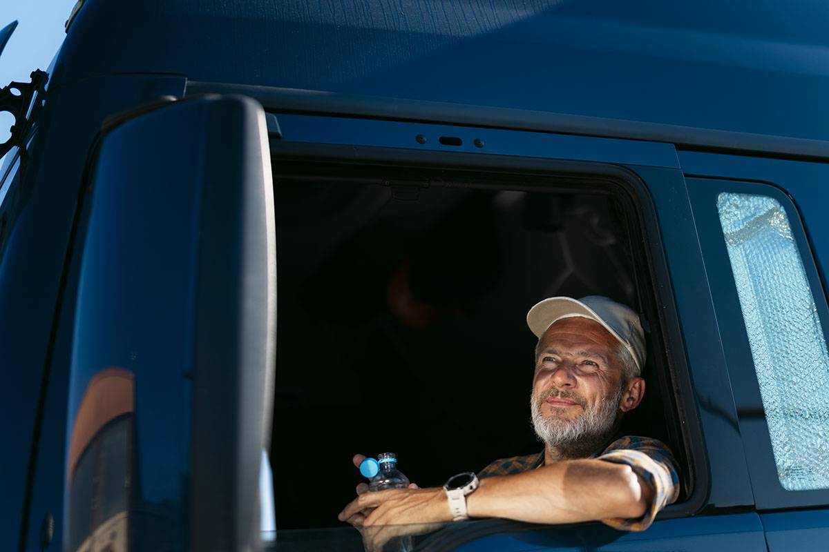 A truck driver in a truck cab window holding an open bottle of water.