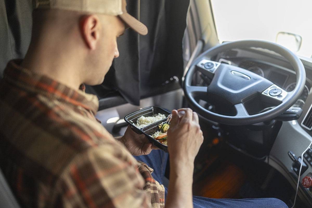 A truck driver sits in a truck cab eating veggies and rice from a container.