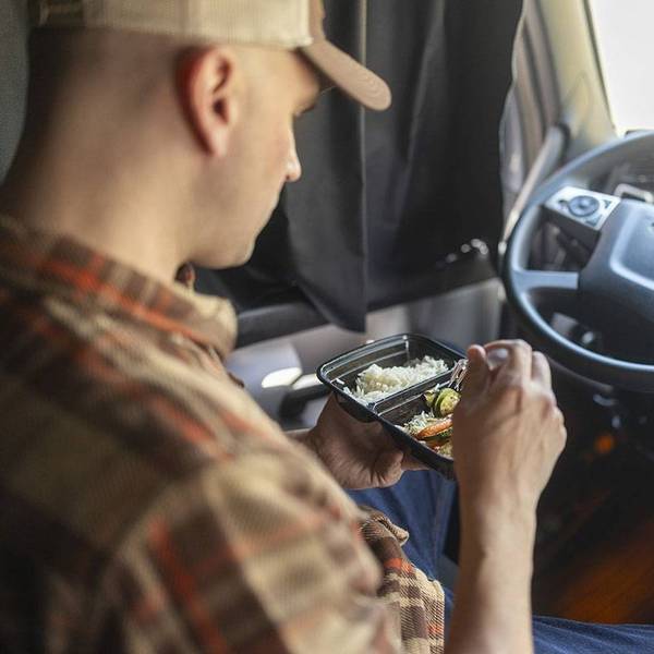 A truck driver sits in a truck cab eating veggies and rice from a container.