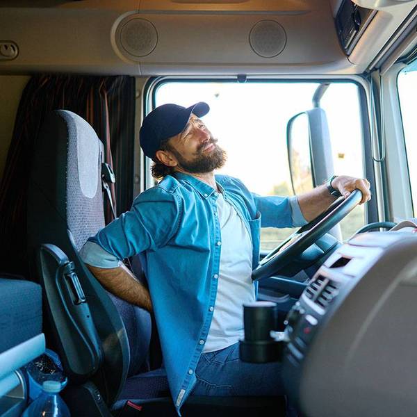 A truck driver sits in the cab of a semi-truck arching his back in pain.