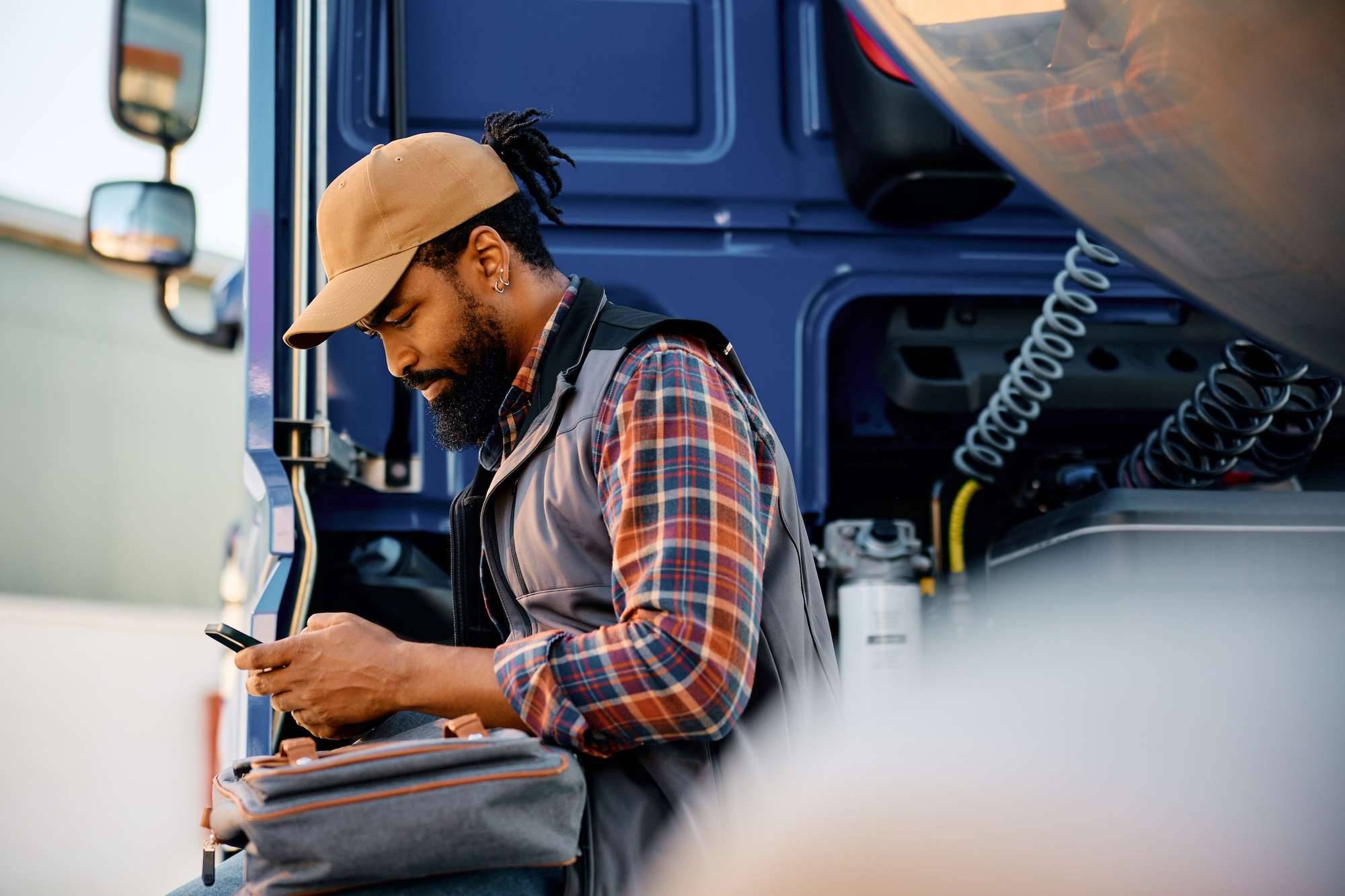 A truck driver sits on the side of his semi-truck while looking at a handheld device.