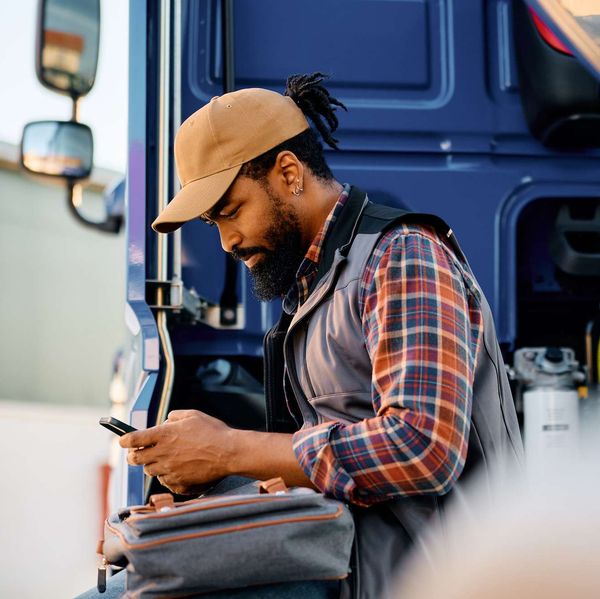 A truck driver sits on the side of his semi-truck while looking at a handheld device.