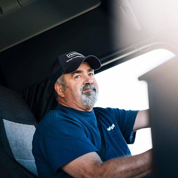 A truck driver with a beard and blue Penske baseball hat sitting in the seat of a truck.