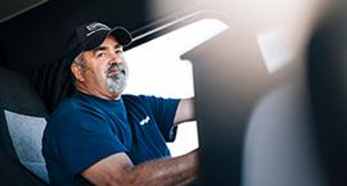 A truck driver with a beard and blue Penske baseball hat sitting in the seat of a truck.